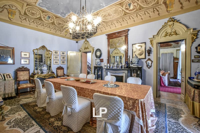 Ornate and luxurious dining room with a large chandelier, intricate ceiling details, antique furniture, and a fireplace. The room has a formal and elegant atmosphere.