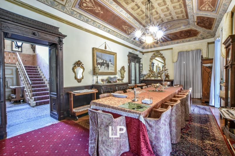 Ornate and elegant dining room with a large wooden table, ornate chairs, a fireplace, and decorative wall hangings and mirrors. The room features a detailed and intricate ceiling design with chandeliers. The overall style is traditional and luxurious.