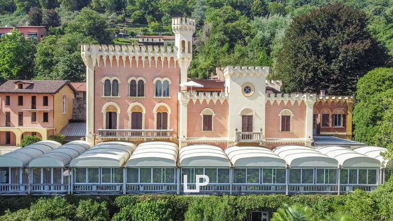 A colorful, ornate castle-like building with arched windows, balconies, and towers surrounded by lush greenery. The building appears to be a hotel or resort with a row of covered outdoor seating areas in front.