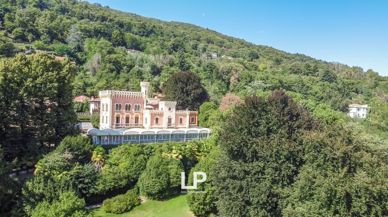 A large, ornate castle-like building surrounded by lush, green vegetation and trees. The building has multiple floors, arched windows, and a balcony or terrace. The property is situated in a scenic, mountainous area.