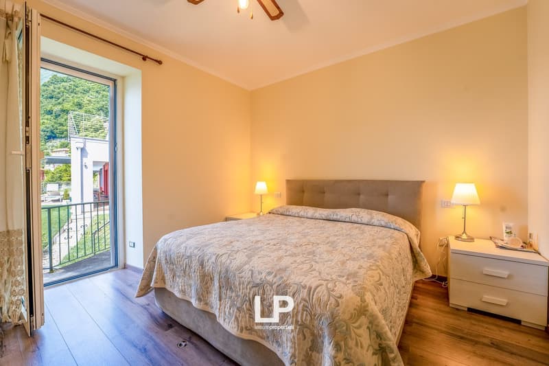 Spacious bedroom with a large bed, bedside tables, and a balcony overlooking the surrounding greenery. The room has warm, neutral-toned walls and hardwood floors.
