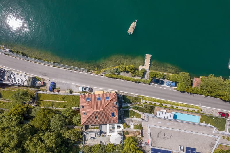 Aerial view of a multi-story residential building with a red tiled roof, surrounded by lush greenery and overlooking a body of water. There is a swimming pool visible on the property, and the building appears to have balconies or terraces. The location of