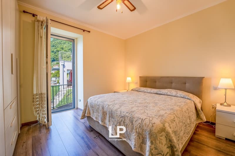 Spacious bedroom with hardwood floors, large window with balcony access, tufted headboard, and a ceiling fan light fixture.