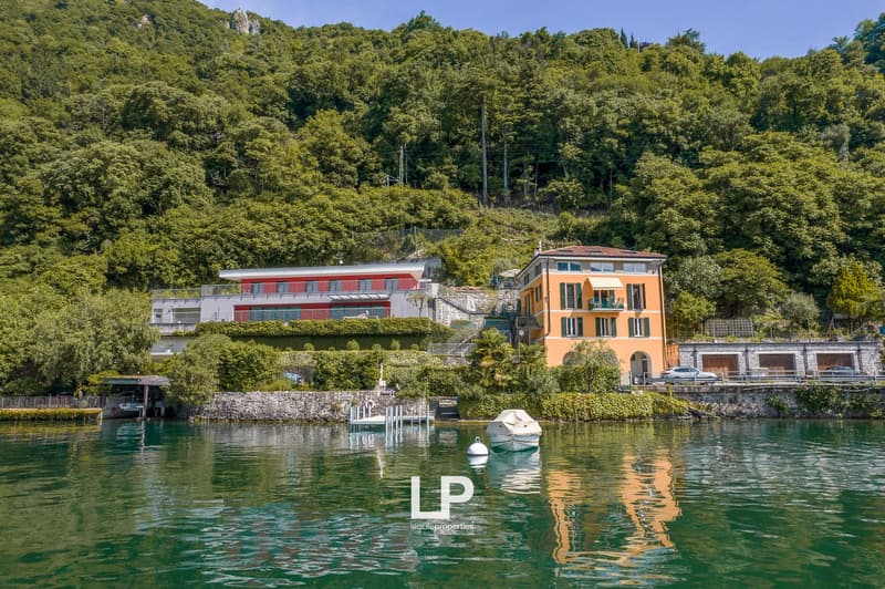 Two-story building with orange and red exterior, surrounded by lush green trees and vegetation, located on the waterfront with a boat dock and parking area