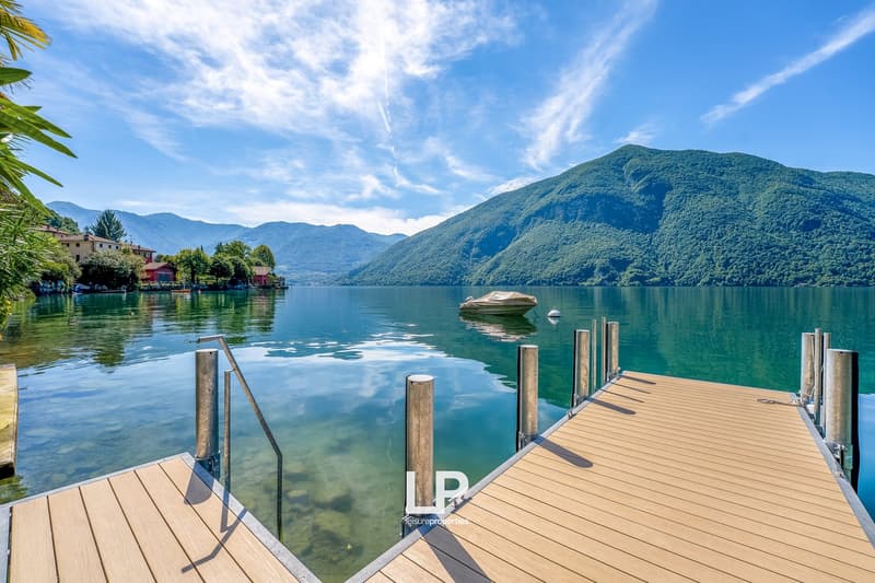 Scenic lakeside view with a wooden dock, a small boat, and mountains in the background. The water is calm and reflects the surrounding landscape. Palm trees and other greenery are visible along the shore.