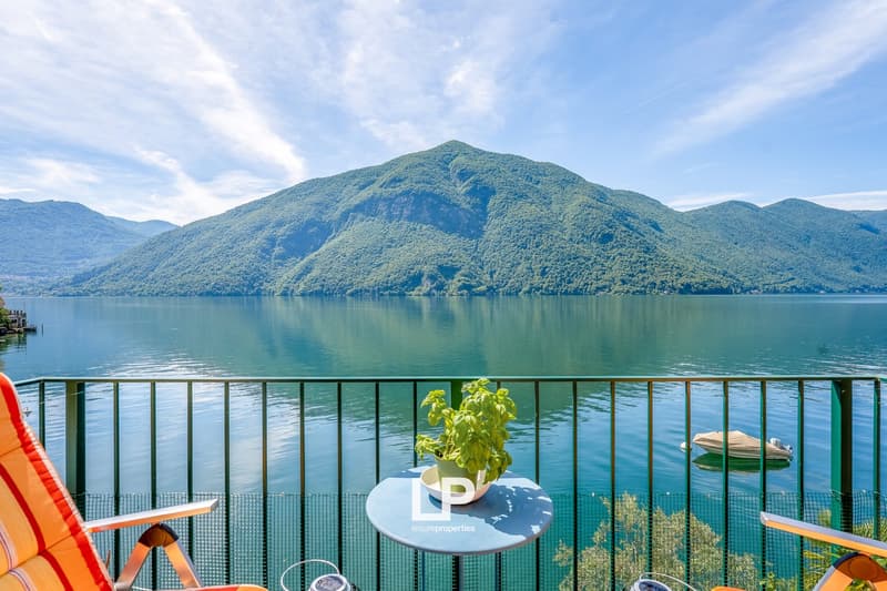 The image shows a lakefront property with a balcony or terrace overlooking a scenic lake surrounded by lush, green mountains. The balcony has a small table with a potted plant, and there is a boat visible on the calm, blue waters of the lake. The property