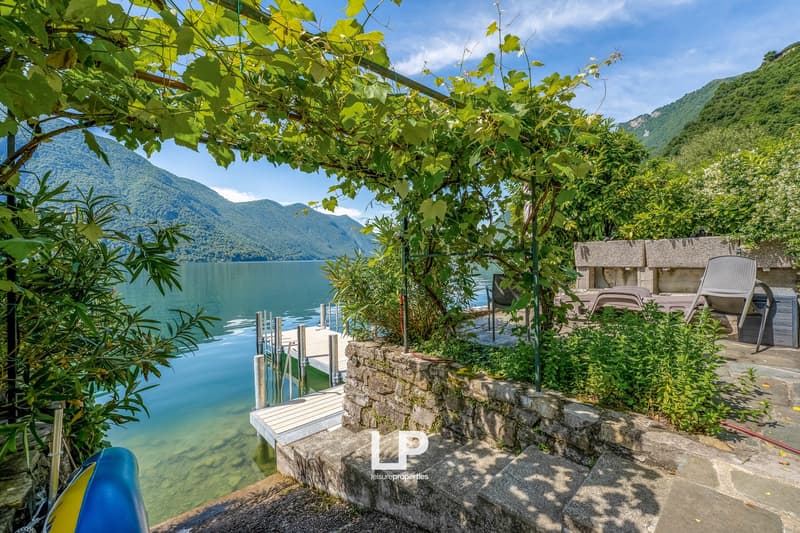Scenic lakeside setting with lush greenery, stone walls, wooden deck, and outdoor furniture overlooking a tranquil lake surrounded by mountains