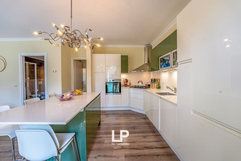 Modern kitchen with white cabinets, stainless steel appliances including a dishwasher, a large island with a dining table, and a chandelier overhead.