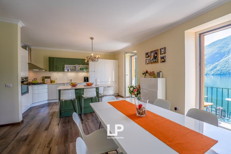 Open-concept kitchen and dining area with large windows overlooking a scenic lake or body of water. The kitchen features white cabinets, green accents, and modern appliances. The dining table has an orange tablecloth and is surrounded by white chairs. The