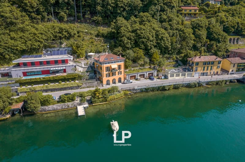 Lakeside buildings with colorful facades, surrounded by lush greenery and a clear blue lake. The buildings appear to be residential or commercial properties, with some having balconies or terraces overlooking the scenic landscape.