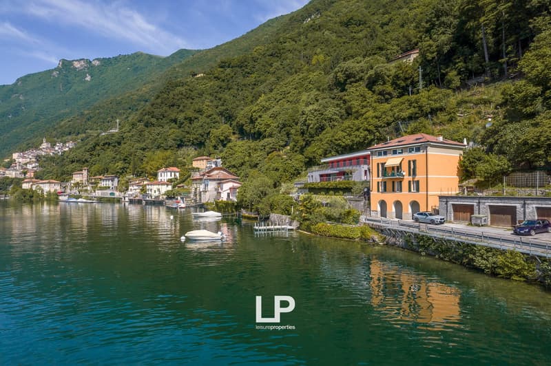 lakefront houses on hillside, greenery, mountain in the background, parked cars, boats, calm water