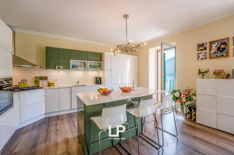 Spacious open-plan kitchen with green and white cabinets, kitchen island with bar stools, hardwood floors, large windows with a view, chandelier lighting, and decorative elements like vases and framed artwork.