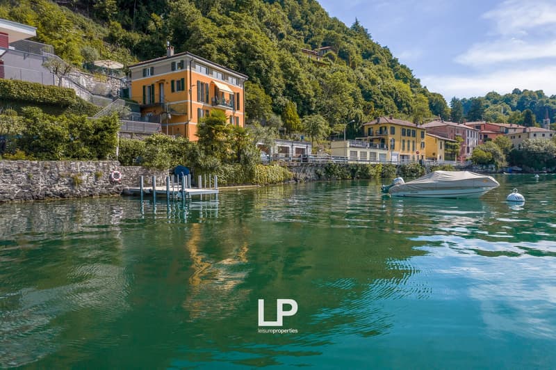 Colorful multi-story buildings with balconies overlooking a scenic lake, surrounded by lush greenery and mountains. A boat is docked in the foreground on the calm, turquoise-colored water.