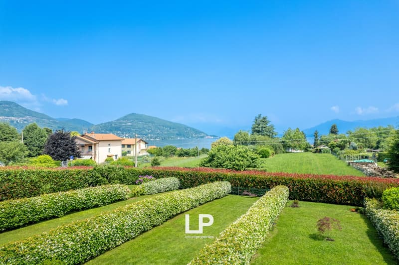 The image shows a scenic landscape with a residential area surrounded by lush greenery, including well-manicured gardens, hedges, and trees. The homes appear to be situated on a hillside overlooking a body of water, likely a lake or bay, with mountains in