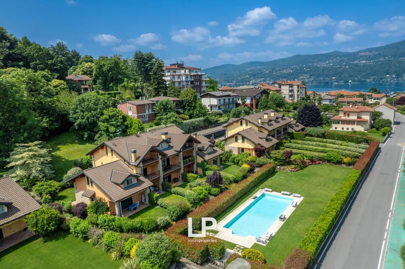The image shows a scenic residential area with a mix of multi-story apartment buildings and single-family homes surrounded by lush greenery and landscaping. The properties have well-maintained gardens and a swimming pool is visible in the foreground. The
