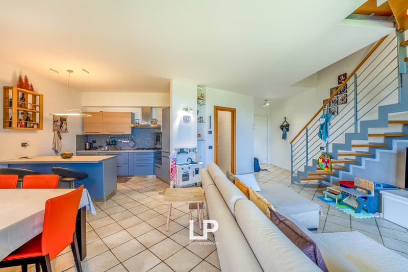 Open-concept living space with kitchen, living room, and dining area. Hardwood floors, modern kitchen with blue cabinets and tile backsplash, dining table with red chairs, sofa, and staircase leading to upper level.