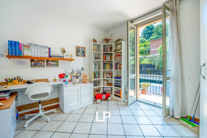Spacious home office with large windows, bookshelves, desk, and various office supplies and decor items. The room has tile flooring and opens up to a balcony or terrace area.