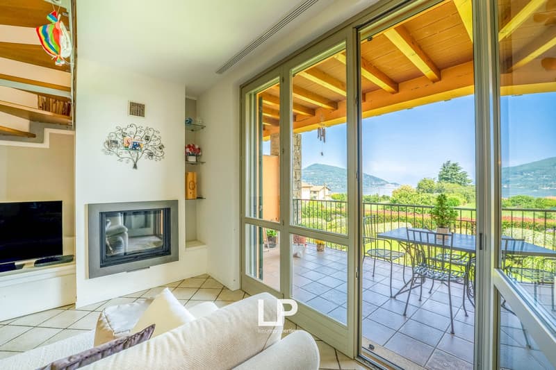 Spacious living room with large windows and sliding glass doors leading to a balcony or terrace, offering a scenic view of the surrounding landscape. The room features a fireplace, wooden beamed ceiling, and tiled flooring.