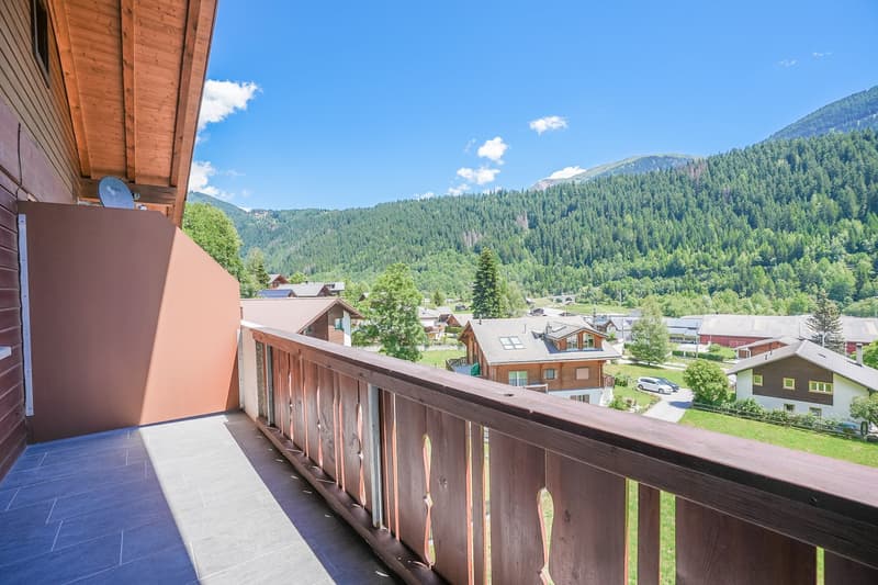 Wooden balcony with a view of a mountainous, forested landscape. The balcony overlooks a small town with various buildings and houses surrounded by lush greenery.