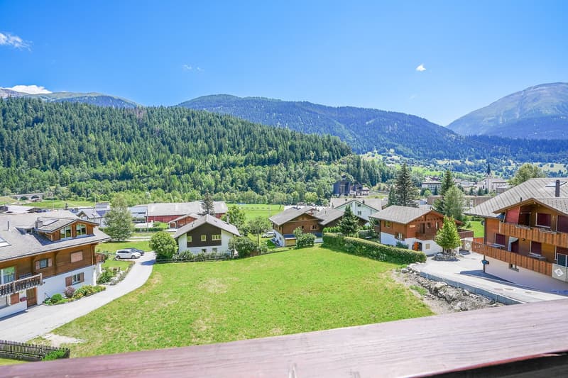 The image shows a scenic mountain landscape with a small town nestled in the valley. The town consists of traditional alpine-style houses with wooden exteriors and sloped roofs. There are grassy fields and lush green forests covering the surrounding hills