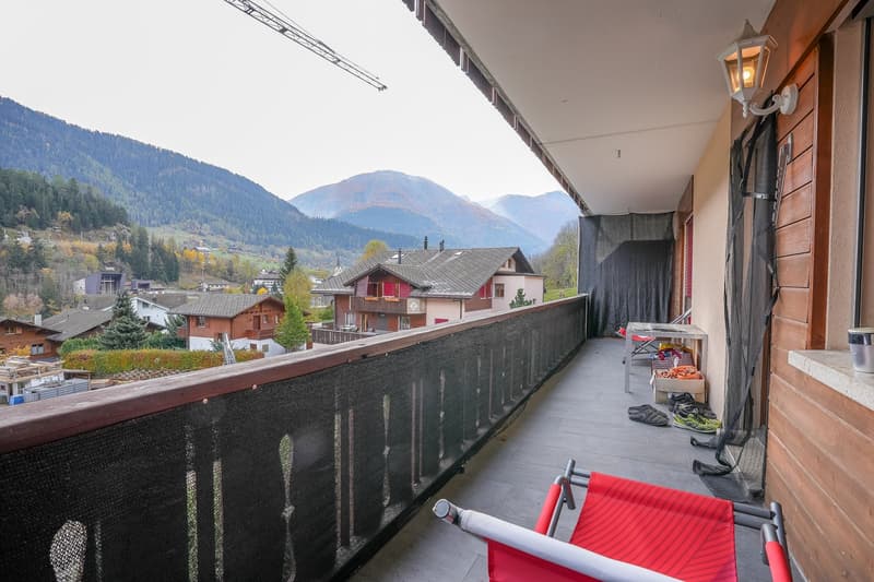 Balcony with a view of a mountainous landscape, wooden houses, and a ski lift in the distance. The balcony has a red chair and some outdoor equipment visible.