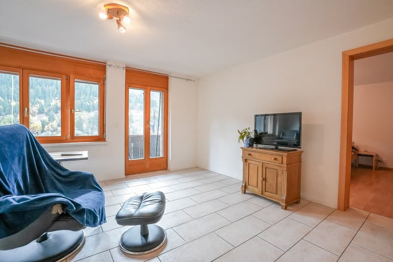 Spacious living room with large windows, wooden frames, tiled floor, sofa, TV stand, and a balcony or terrace with a view of the surrounding forest