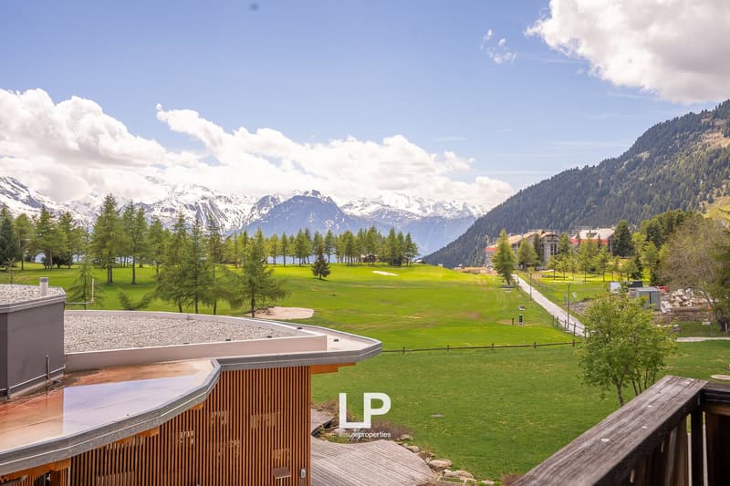 The image shows a scenic mountain landscape with a large grassy field or golf course in the foreground. The field is surrounded by tall pine trees and the background features snow-capped mountains. There are several buildings or structures visible, includ