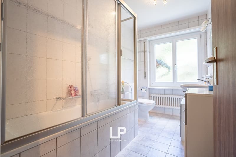 Spacious bathroom with tiled walls and floors, large shower enclosure, toilet, and vanity sink. The room has a bright and clean appearance with a window providing natural light.