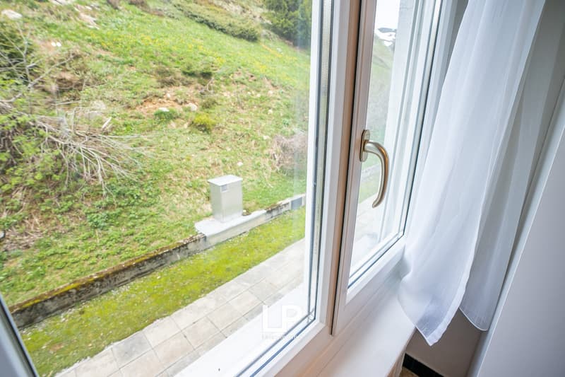Large window with a view of a grassy, hilly landscape outside. The window has a wooden frame and a metal handle. The floor inside appears to be tiled.