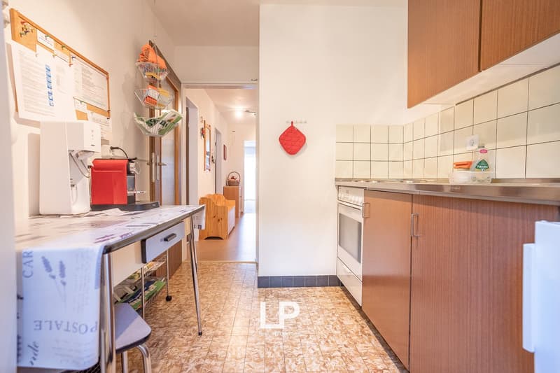 Galley-style kitchen with white tile walls, wooden cabinets, stove, dishwasher, and washing machine. Shelving unit and various kitchen items visible. Tiled floor and hallway leading to other rooms.