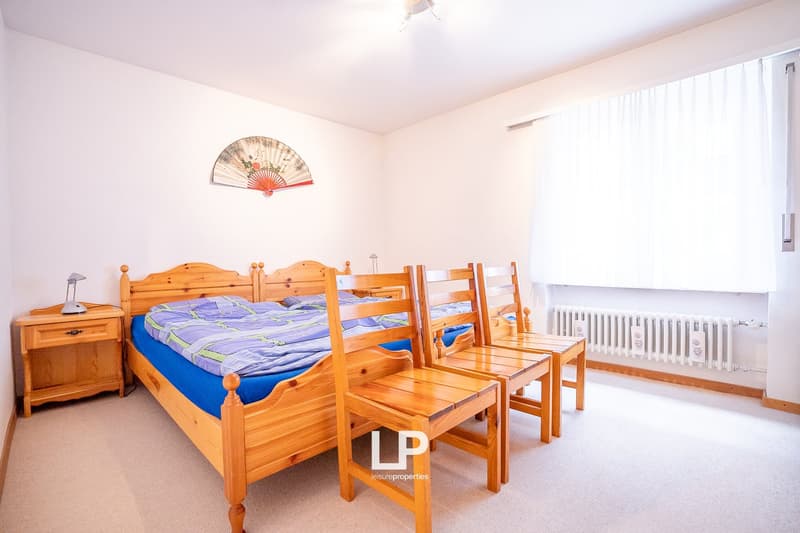Two wooden beds, wooden night stands with lamps, wooden chairs, wooden floor, white walls, white radiator, window with white curtains, decorative fan on wall