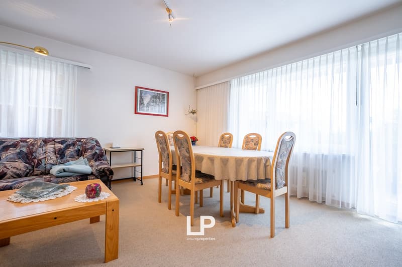 Spacious living room with a dining area, wooden furniture including a table and chairs, a sofa, and large windows providing natural light.
