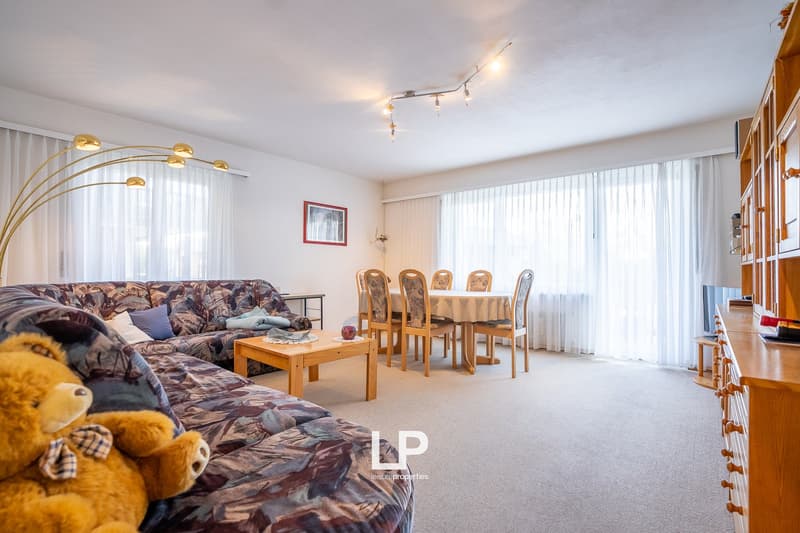 Spacious living room with a large patterned sofa, wooden coffee table, and dining table with chairs. The room has large windows providing natural light and a track lighting system. There is also a bookshelf visible on the side.