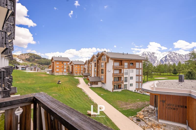 Wooden buildings, well-kept grass, pathway, mountains in the background, balcony railing, clear blue sky