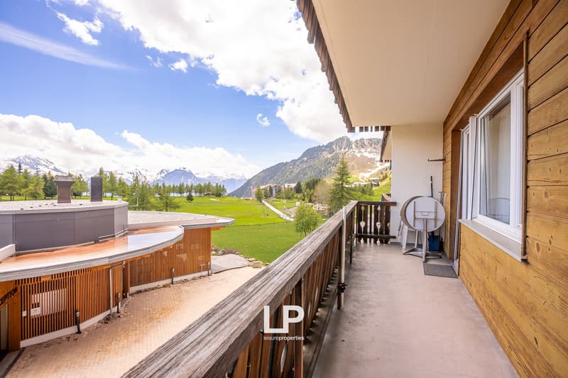 Wooden balcony with a view of the mountains and green landscape, surrounded by pine trees. The apartment has large windows and a wooden exterior design.