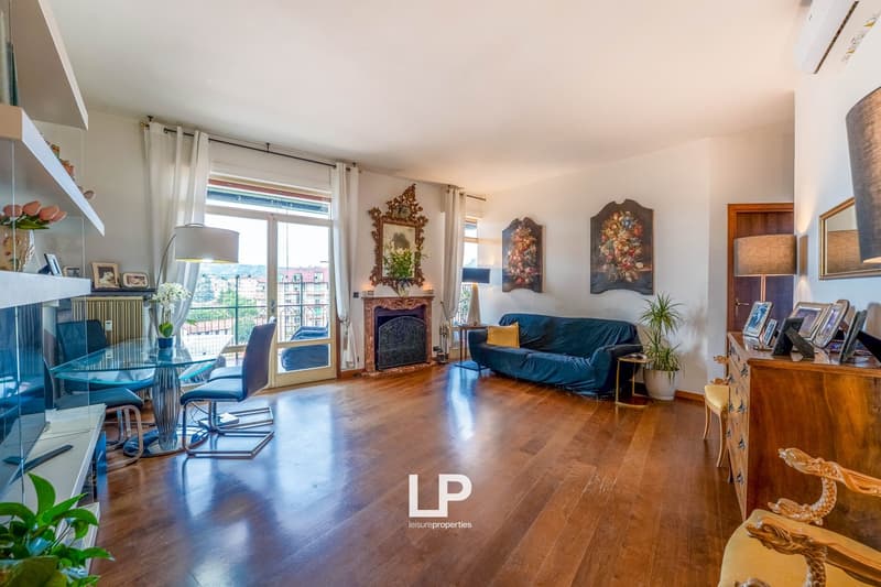 Spacious living room with large windows, hardwood floors, blue velvet sofa, antique-style decor, and a balcony or terrace overlooking the city