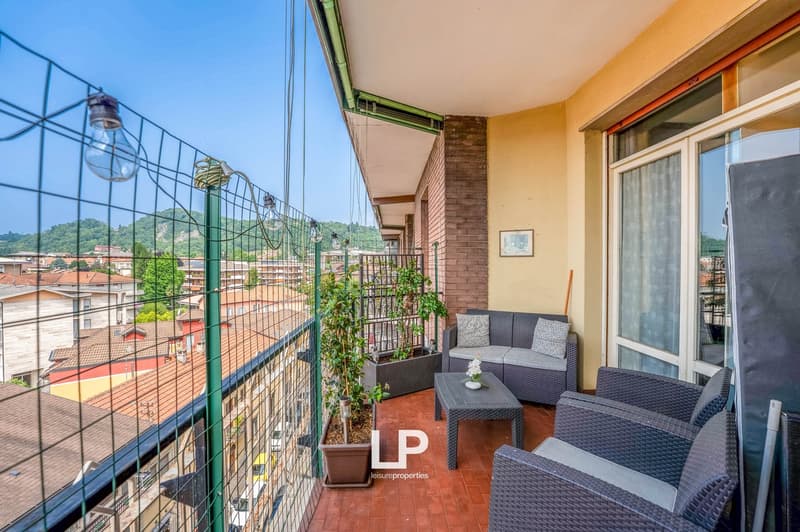 Balcony with outdoor furniture, potted plants, and a view of the surrounding buildings and greenery. The balcony has a brick wall and metal railing.
