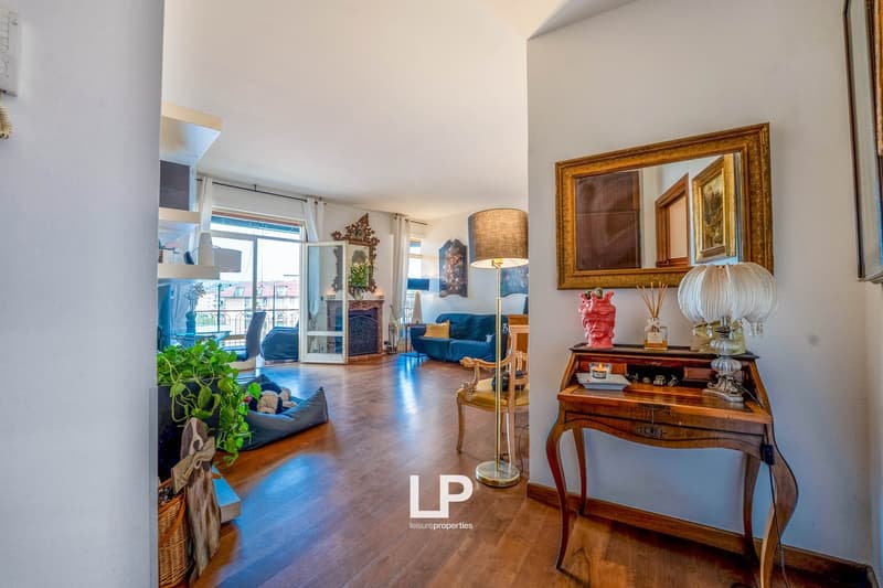 Spacious living room with hardwood floors, white walls, large windows, blue sofa, wooden console table with decorative items, and a balcony visible through the windows.