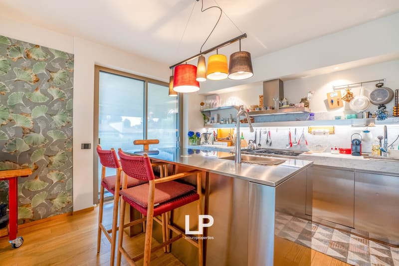 Modern kitchen with stainless steel appliances, open shelving, colorful pendant lights, and a breakfast bar with red stools. The kitchen has a sliding glass door leading to a balcony or terrace.