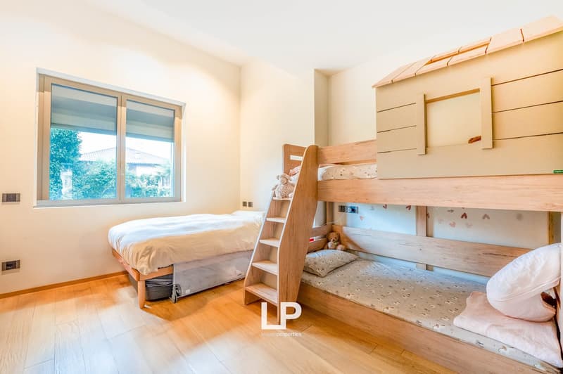 Spacious bedroom with wooden bunk beds, a single bed, and large windows providing natural light. The room has a warm, cozy atmosphere with wooden floors and neutral-colored walls.