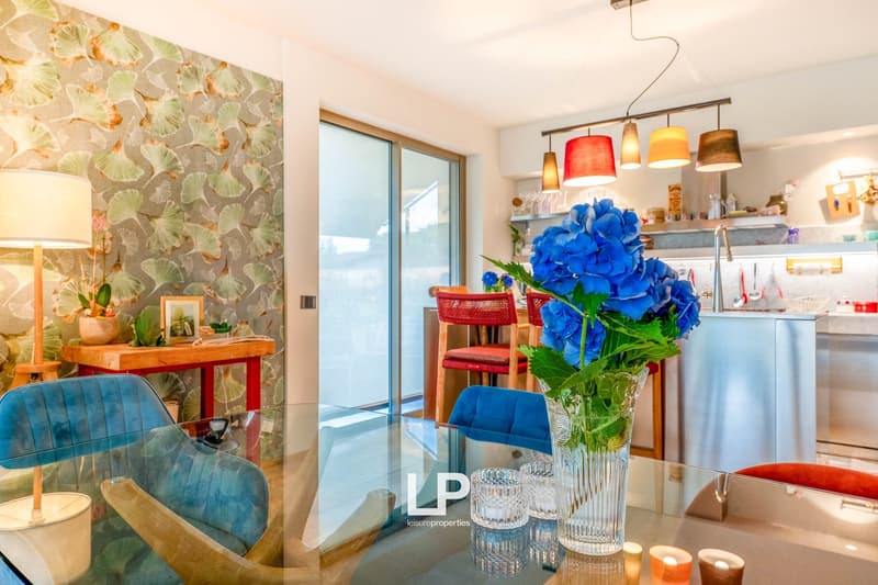 Open kitchen with large windows, blue and red accents, floral wallpaper, dining table with red chairs, blue sofa, and vase of blue flowers on the table