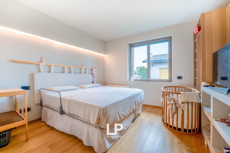 Spacious bedroom with large windows, wooden flooring, a king-size bed, a crib, and wooden furniture including a dresser and shelves.