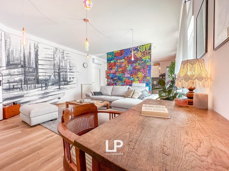 Spacious living room with hardwood floors, white walls, large windows, colorful graffiti-style artwork, gray sofa, wooden coffee table, and decorative lamps