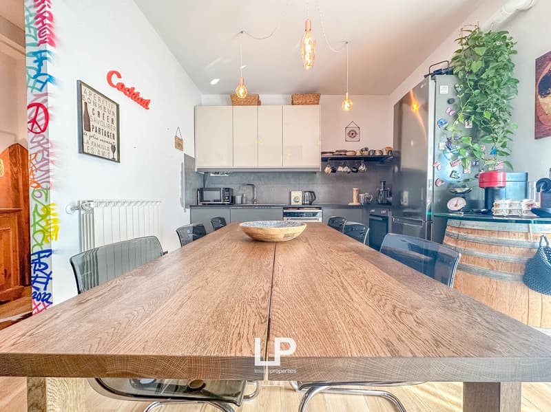 Large wooden dining table, modern kitchen with white cabinets, black appliances, hanging light fixtures, and a potted plant