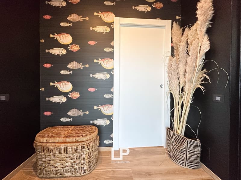The image shows a room with a white door, a wicker basket, and a vase filled with dried plants. The walls are decorated with a wallpaper featuring a pattern of colorful fish.
