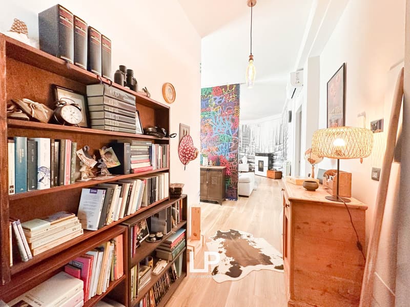 Wooden bookshelves with various books and decor items, hardwood floors, colorful artwork on the wall, a wooden cabinet with lamps, and a cozy living space with a fireplace visible.