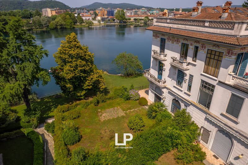 Ornate historic building with balconies overlooking a lake, surrounded by lush greenery and trees. The building has a traditional architectural style with tiled roofs. The cityscape in the background includes modern high-rise buildings.