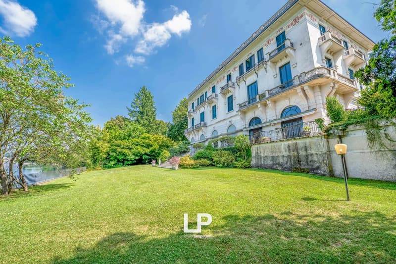 A large, multi-story building with a classical architectural style, featuring balconies and surrounded by a well-manicured lawn and lush greenery.