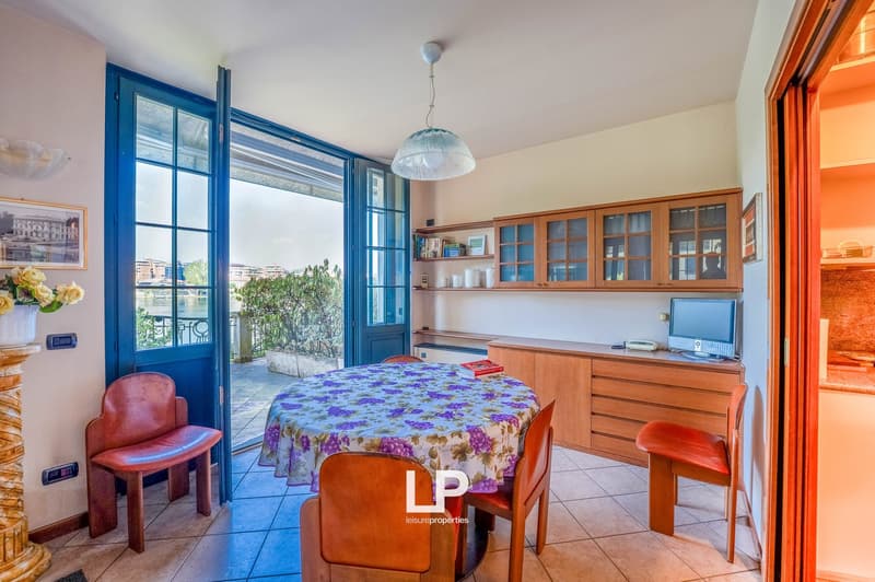 Spacious kitchen and dining area with large windows overlooking a balcony or terrace, wooden cabinets and shelves, tiled flooring, dining table with floral tablecloth and chairs, and a pendant light fixture.