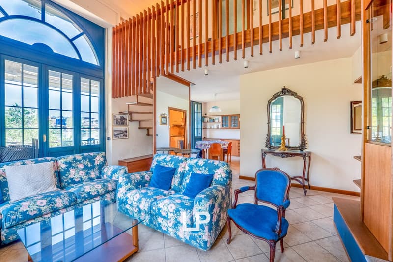Spacious living room with large arched windows, wooden ceiling beams, floral patterned sofas, antique-style furniture, and a decorative mirror on the wall